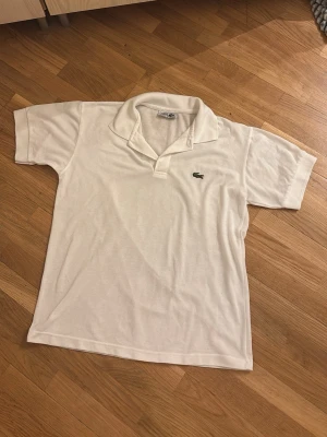 Vit kortärmad t-shirt från Lacoste - Klassisk vit t-shirt från Lacoste med korta ärmar och ikoniska krokodilloggan på bröstet. Tillverkad i mjuk bomull och har en snygg krage med v-ringad öppning. Perfekt för en clean och stilren look.