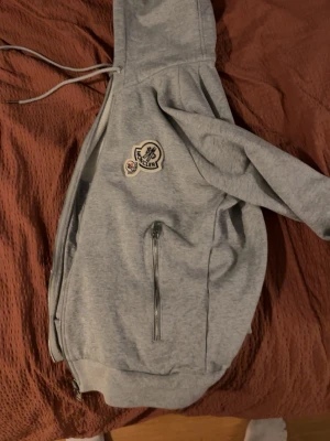 Grå moncler hoodie - Snygg grå hoodie med dragkedja, huva med snören och en patch på bröstet. Mjuk insida och praktiska fickor med dragkedja. Perfekt för chill dagar eller när du vill ha en enkel streetstyle-look.