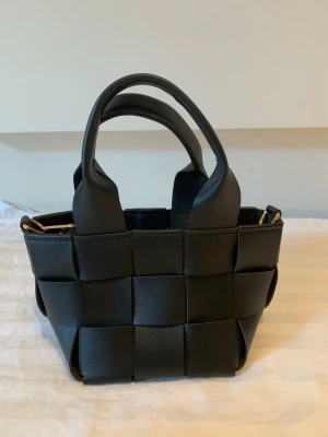 Svart flätad minihandväska - Trendig svart minihandväska i flätad design med både korta handtag och avtagbar axelrem. Väskan är tillverkad i ett slätt, konstläderliknande material och har guldfärgade metalldetaljer. Perfekt för dig som gillar stilrena accessoarer med en modern twist.