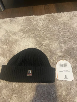 Svart ribbstickad mössa Parajumpers - Snygg svart ribbstickad mössa från Parajumpers med uppvikt kant och klassisk logga framtill. Mössan har en diskret etikett på insidan och är tillverkad i mjukt material som håller dig varm under kalla dagar.
