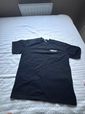Svart Essentials Fear of God t-shirt - Säljer en svart t-shirt från Fear of God Essentials med tryck på bröstet och stor logga på ryggen. Klassisk passform och korta ärmar. Perfekt för dig som gillar streetwear och vill ha en enkel men snygg look.