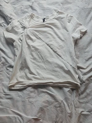 Vit t-shirt med draperad detalj - Säljer en vit t-shirt i bomull med korta ärmar och unik draperad detalj framtill som ger en cool twist till den klassiska looken. Perfekt för dig som vill ha något enkelt men ändå speciellt i garderoben.