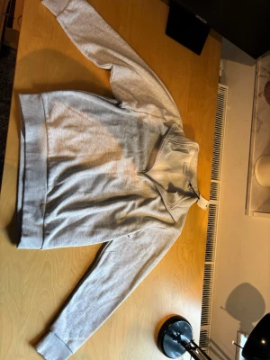 Quarter zip från Jack and Jones  - En stilren ljusgrå hoodie i mjukt sweatshirtmaterial. Hoodien har långa ärmar, ribbade muddar och bred fåll nedtill. Klassisk passform utan tryck eller mönster, perfekt för en avslappnad look. Ingen synlig logga eller märke.