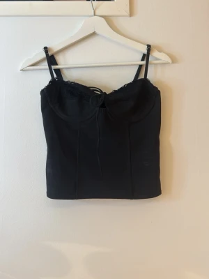 Svart korsettopp med spetsdetaljer - Snygg svart korsettopp med tunna axelband och spetsdetaljer upptill. Toppen har snörning framtill och dragkedja bak för en tight passform. Perfekt att styla med höga jeans eller kjol för en trendig look.
