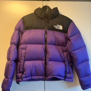 The North Face - En lila/svart dunjacka från The North Face. Jackan är 1år gammal. Har en luva som går att stoppa in i kragen, 1 innerficka med dragkedja och en fick på varje sida som stängs med dragkedja. Jackan har dragsko för önskad passform nertill. Storlek M