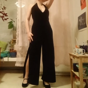 Jumpsuit med slits - Svart jumpsuit i sammetsmaterial med djup ringning och slits på båda benen. Halterneck, knäpps i nacken med två knappar. Öppen rygg. Lapp är bortklippt men gissar på att den är storlek 36-38. Stretchigt tyg.