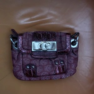 Väska Guess - Lila vintage guessväska i monogram! Lite missfärgad framtill på loggan i silver samt att axelbandet har lite skavanker!