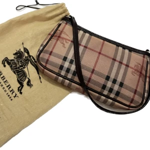 Burberry väska - Säljer denna burburry väskan/plånbok som man kan ha som axelväska som passar till allt. Självklart ÄKTA. Inuti är det ett litet fack som är perfekt till att ha kort i. Har dustbagen men tyvärr inte något tillbehör kvar. Mått: 23x13x2,5 cm.