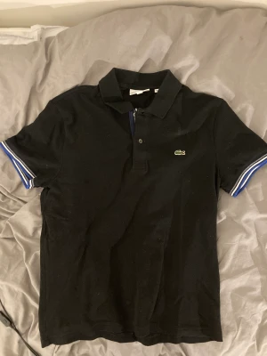 Svart lacoste piké - Svart lacoste piké med blåa/vita strämg på ärmarna  Storlek M   Möts görna upp i malmö annars står du för frakten🤡