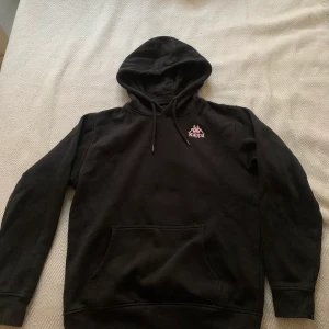 Hoodie Kappa - En svart hoodie av märket Kappa. Det sitter en liten rosa broderad logga på framsidan samt en stor tryckt logga på ryggen. Den är i strl S och är normal i storleken. Sparsamt använd. 