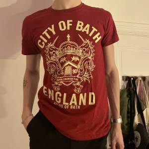 Röd graphic tee köpt på second hand med ett ascoolt gult stort tryck av ett emblem och texten ”City of Bath, England” ❤️⚜️