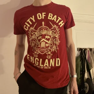 Röd graphic tee med gult stort tryck emblem - Röd graphic tee köpt på second hand med ett ascoolt gult stort tryck av ett emblem och texten ”City of Bath, England” ❤️⚜️