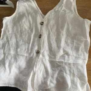 Vitt linne - Oanvänd vitt topp med volang nedtill med bruna knappar.  Material- linnetyg ingen stretch i tyget så liten i storleken. Storlek 34 men drar sig mer åt 32 tycker jag. Är helt oanvänd bara provad! (Köparen står för den eventuella fraktkostnaden)
