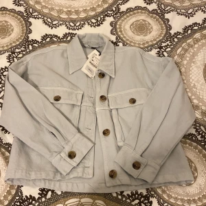 Kort skjortjacka i jeans från Zara i storlek S  - Oanvänd jeansjacka med prislapp från Zara säljes. Har haft jackan i några månader men inte använt den så det är dags att den får en ny ägare😊. Färgen är ljusblå med något grått inslag (syns på sista bilden)😇