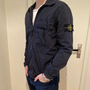 Stone island overshirt - Säljer en Stone island overshirt i färgen mörkblå som är en riktig klassiker till sommaren! Overshirten har använts men varit väl omhändertagen och därför inga större defekter. Skulle säga att overshirten är 9/10. Äkta såklart. Stone island påse finns med