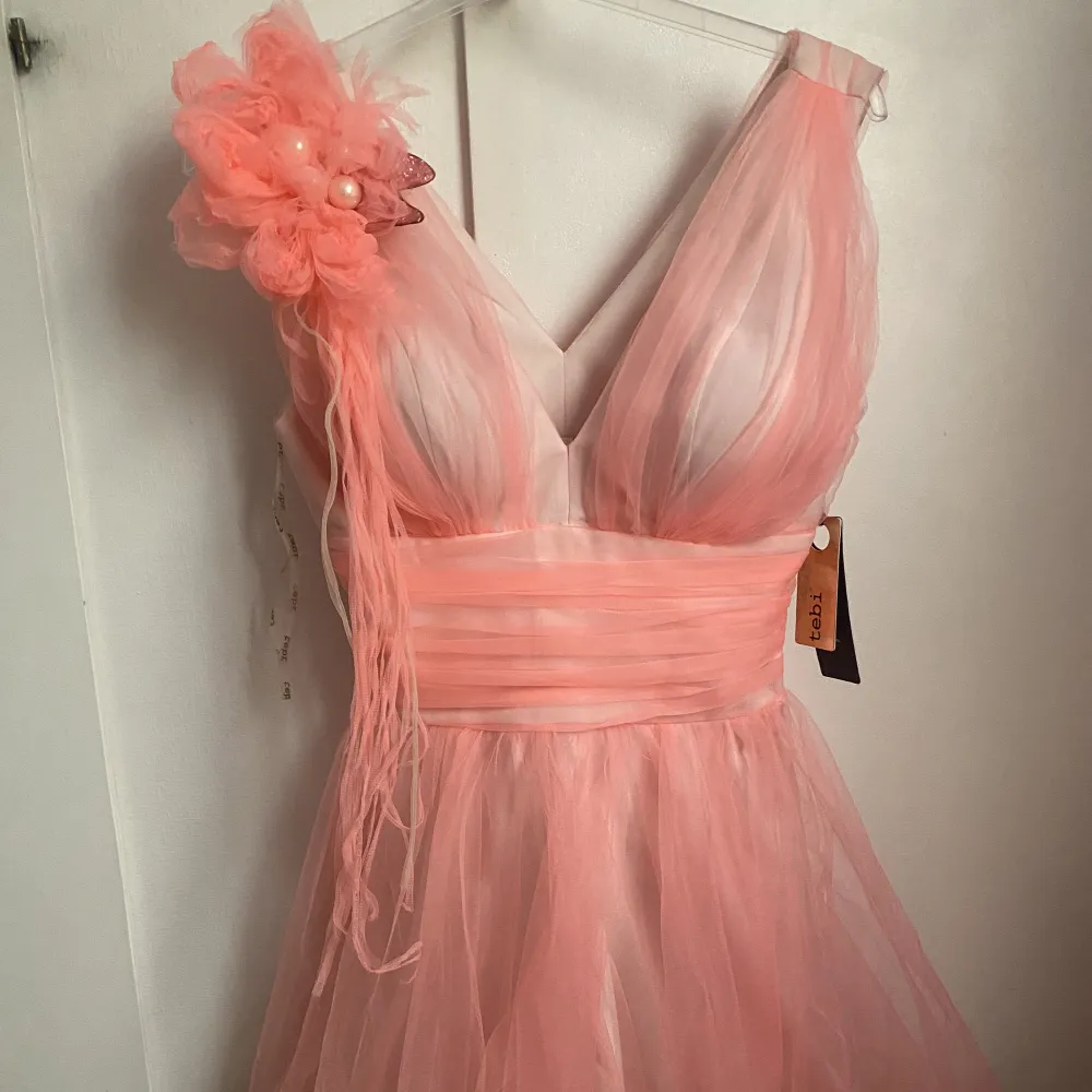 Corall/rosa fest klänning som är ungefär knälängd. Oanvänd med lapp på. ”Rosen” är justerbar så man kan lägga den på valfri plats eller ta bort den.  Passar till större fester som bröllop, födelsedag etc.  . Klänningar.