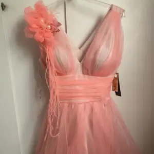 Corall/rosa fest klänning som är ungefär knälängd. Oanvänd med lapp på. ”Rosen” är justerbar så man kan lägga den på valfri plats eller ta bort den.  Passar till större fester som bröllop, födelsedag etc.  
