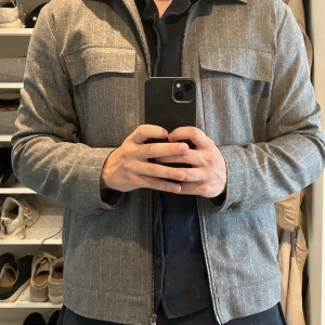 Overshirt - Overshirt från J.Lindeberg från deras FW22 kollektion. Plagget är grått med beiga tunna linjer i som är riktigt snygga detaljer till jackan. Den är använd sparsamt så iprincip ny. Storlek M men skulle även funka på en S