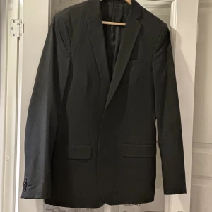 Kostym  - En stilig svart kostym med väldigt tunna gråa linjer i stl 50. Använd endast en gång. Kostymen består av kavaj och slimmad byxa. Mycket elegant. 