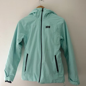 Skidjacka Helly Hansen - Jättefin skidjacka från Helly Hansen. Använd 1 vecka i alperna. Den är fodrad, har snölås, luftfickor under armhålorna och har liftkortsficka på armen. Går att ha hjälm med luvan. Finns lite slitage på vänster underarm. Fin blå färg som syns i backen!⛷️