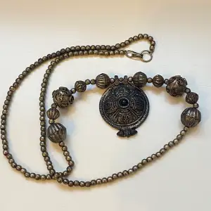 Vackert rustikt halsband med tyngd i hänget  Berlocken är utsmyckad med vackert mönster och svart sten   Okänt material 