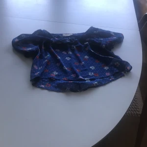 Blus - En blå blus med gröna prickar och vita och röda blommor. Är lite genomskinlig men är bra ändå! Kostar 150 kr! 