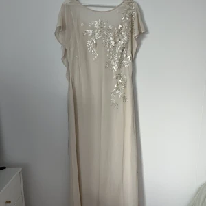 Klänning - Jättefin Vit/Beige långklänning. Aldrig använd, med prislappen kvar. Med detaljer på. Öppen rygg med en fin knytning. Passar till Bröllop, bal m.m