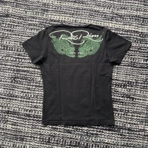 T-shirt med tryck - Ed hardy liknande t-shirt med coolt tryck både på bröstet och ryggen! Köpt second hand men sparsamt använt av mig och har inga defekter. Står ingen storlek men skulle gissa på M. Kontakta mig med frågor/intresse!!