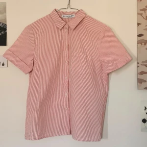 Rodebjer Hathaway Skjorta kortärmad - Kortärmad, randig skjorta i bäckebölja. Vit och röd. Uppvikta ärmslut. 100% bomull, kraftig kvalitet.  Längd 55 cm. 