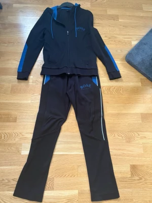 Svarta mjukisbyxor från BOSS och tröja - Mysigt svart loungewear-set från BOSS med blå detaljer. Setet består av mjukisbyxor med blå logga och matchande hoodie med dragkedja, blå huva och blå ränder längs ärmarna. Tillverkat i mjukt material för maximal komfort.