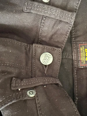 Svarta jeans från Lee - Säljer ett par svarta jeans från Lee med klassisk femficksdesign och raka ben. Jeansen har metallknappar med Lee-logga och är tillverkade i ett slitstarkt bomullsmaterial. Perfekta för en stilren och enkel look.