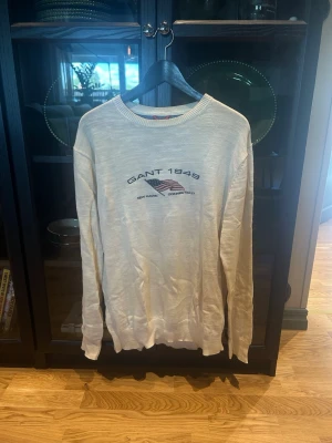Beige/vit tröja från Gant med tryck - Snygg beige tröja från Gant med tryck på bröstet där det står 'GANT 1949' och en amerikansk flagga. Tillverkad i 100% bomull och har rund halsringning samt långa ärmar. Perfekt för en avslappnad och stilren look.