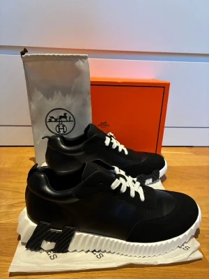 H é r m e s sneakers  - Snygga svarta H é r m e s sneakers med kontrasterande vita skosnören och en chunky, räfflad vit sula. Skorna har en låg profil och är tillverkade i en mix av läder och textil. Sidan pryds av en svart H e r m é s-logga i 3D. Perfekta för dig som gillar exklusiv streetstyle.