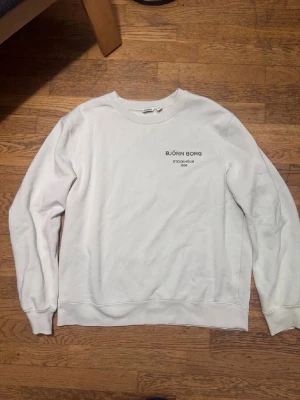 Vit sweatshirt från Björn Borg  - Klassisk vit sweatshirt från Björn Borg med tryck på bröstet där det står 'BJÖRN BORG STOCKHOLM 1956'. Tröjan har rund halsringning, långa ärmar och ribbade muddar vid ärmslut och nederkant. Perfekt för en clean och enkel stil.