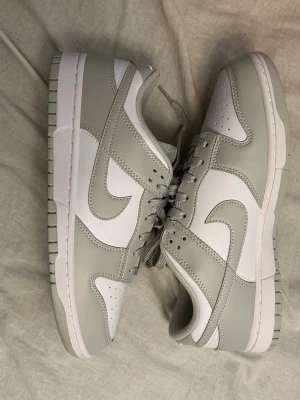 Nike Dunk Low gråvita sneakers - Nike Dunk Low sneakers i stilren grå och vit färg. Skorna har klassisk låg siluett, perforerad tåbox och ikoniska Swoosh-loggan på sidan. Ovandelen är i slätt läder med matchande grå snören och yttersula i gummi. Perfekta för en clean och modern look.