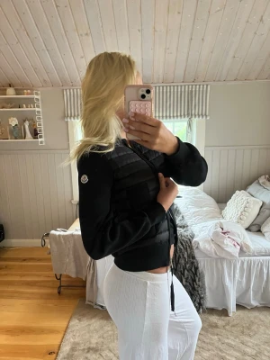 Svart pufferjacka med dragkedja - Snygg svart pufferjacka med quiltade partier och dragkedja framtill. Jackan har en croppad passform och ribbade muddar vid ärmsluten. Perfekt för dig som vill ha en trendig och varm jacka till vintern. Moncler-logga på ärmen för extra stilpoäng.