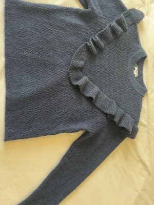 Mörkgrå stickad tröja med volang - Mysig mörkgrå stickad tröja från Gina Tricot med vågigt mönster och en stor volangdetalj som går diagonalt över framsidan. Tröjan har långa ärmar och rund halsringning. Perfekt för dig som vill ha något både snyggt och bekvämt.