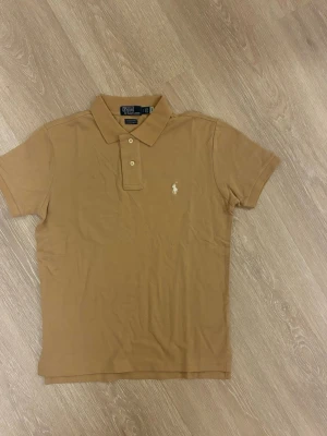 Beige pikétröja från Polo Ralph Lauren - Klassisk beige pikétröja från Polo Ralph Lauren med vit broderad logga på bröstet. Tröjan har korta ärmar, krage och knappar framtill. Perfekt för en clean och stilren look.
