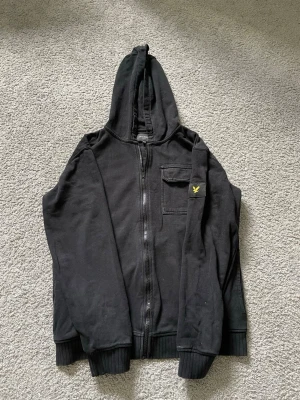Lyle & Scott hoodie  - Säljer en svart Lyle and Scott hoodie med dragkedja och huva. Den har en bröstficka och en gul broderad logga på armen. Långa ärmar och ribbade muddar vid ärmslut och nederkant. Perfekt för chill dagar eller när du vill ha en enkel och clean look. Storlek = 12/13 år.