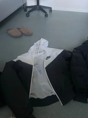 Svartvit vindjacka från Nike - Snygg svartvit vindjacka från Nike med dragkedja och huva. Jackan har en sportig design med meshfoder och logga på bröstet. Perfekt för dig som gillar streetwear och vill ha en lätt jacka till vardags.