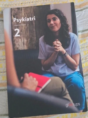 Psykiatri 2 kursbok - Kurslitteratur med titeln 'Psykiatri 2' av Susanne Niemi. Omslaget visar två personer i samtal, där en håller i en röd anteckningsbok. Boken ges ut av Gleerups och har ett modernt, färgfotograferat omslag.