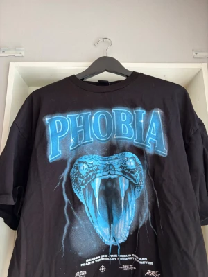 Svart t-shirt med blått ormmotiv - Säljer en svart t-shirt med stort blått tryck av en orm och texten PHOBIA på bröstet. T-shirten har korta ärmar och en relaxed passform. Perfekt för dig som gillar streetwear och grafiska prints.