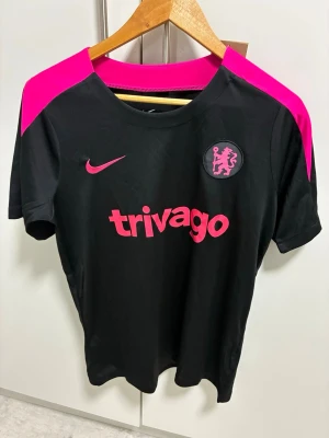 Svart Nike Chelsea tränings T-Shirt - Snygg svart tränings t-shirt från Nike med Chelsea FC-logga och rosa detaljer på axlar, logga och tryck. Tillverkad i ett lätt och snabbtorkande material, perfekt för träning eller match. Trivago som sponsortryck på bröstet och rund hals.