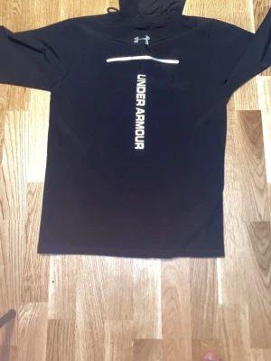 Svart hoodie från Under Armour - Snygg svart hoodie från Under Armour med dragkedja och huva. Stor logga och text framtill i vitt. Tillverkad i ett mjukt och stretchigt material som passar perfekt till både träning och chill. Långa ärmar och minimalistisk design. Pris går att diskuteras🤝🤝