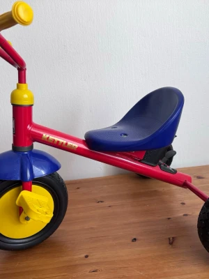 Trehjuling från Kettler i rött och blått - Säljer en klassisk trehjuling från Kettler med röd metallram, blått plastskal på sits och skärm samt gula pedaler och handtag. Trehjulingen har svarta gummihjul och en låg sits som gör den enkel att använda för mindre barn. Perfekt för lek utomhus.