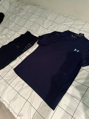 Blå träningsset från Under Armour - Säljer ett blått träningsset från Under Armour med en kortärmad t-shirt och matchande tights. T-shirten har en liten UA-logga i regnbågsfärger på bröstet och tightsen har diskreta logodetaljer. Perfekt för gym eller löpning.