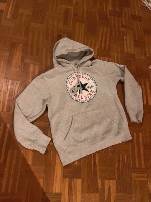 Grå Converse All Star hoodie - Snygg grå hoodie från Converse med det klassiska All Star-logotrycket i vitt, svart och rött på bröstet. Hoodien har huva med dragsko och en stor magficka. Perfekt för en avslappnad streetwear-look. Storlek M sitter som S 