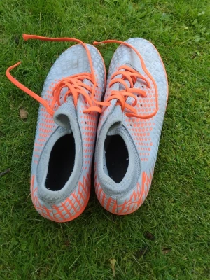 Grå-orange fotbollsskor från Puma  - Snygga fotbollsskor från Puma i grått med orange detaljer och snörning. Skorna har en modern design med mönstrad ovandel och greppvänlig sula i orange. Perfekta för dig som vill sticka ut på planen och få bra kontroll på bollen. Pris kan diskuteras 