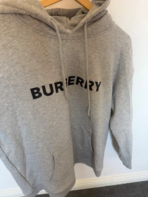 Grå hoodie från Burberry med tryck - Snygg grå hoodie från Burberry med svart logotyptryck över bröstet. Hoodien har en klassisk huva med dragsko och en stor magficka framtill. Tillverkad i mjukt sweatshirtmaterial som är perfekt för en avslappnad stil.