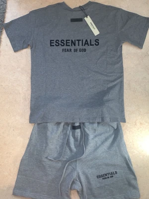 L. Äkta Essentials summer set - Köpte och dom kom hem va då utomlands då har man har 14 dagar retur retlighet va utomlands 20 dagar när jag kom hem så va dom lite tight för mig Säljer ett grått set från Essentials Fear of God med t-shirt och shorts. Setet är gjort i mjukt bomullsmaterial och har svart tryck med loggan på både tröjan och shortsen. Perfekt för en avslappnad och trendig look.